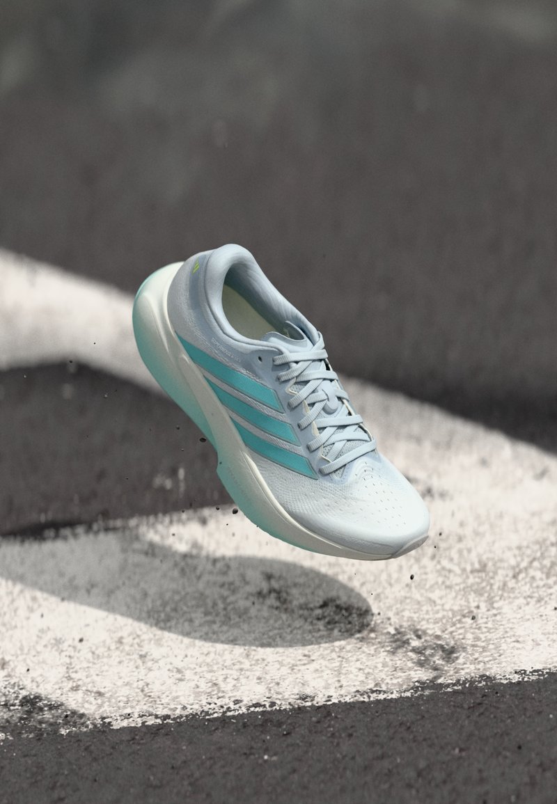 Zapatilla deportiva azul claro con superficie texturizada, tres franjas azul oscuro y suela acolchada, suspendida sobre un fondo de asfalto negro.