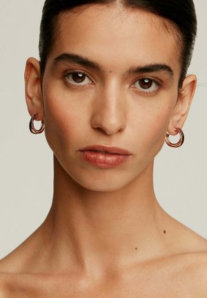 Pendientes de aro en oro rosa con un diseño retorcido, reflejando la luz; textura suave y forma circular. Fondo sutil de tono de piel.