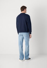 Homem com um sweatshirt azul marinho, jeans claros e ténis pretos, de pé de costas para uma parede branca simples.