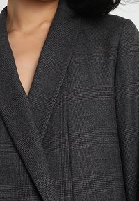 Šedý kostkovaný blazer s klopovým límcem a strukturovanými klopami, vyrobený z texturované tkaniny s jemným kostkovým vzorem.