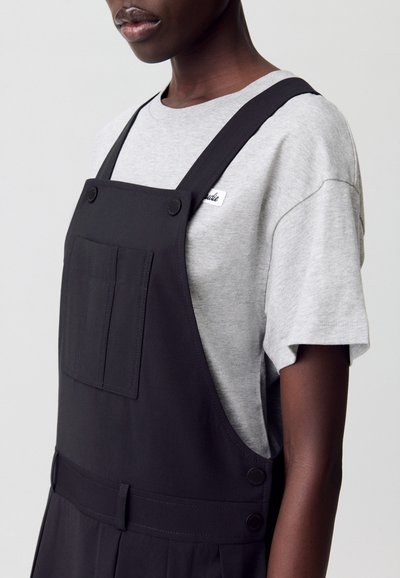 Primer plano de una persona que lleva una camiseta gris debajo de un mono negro, con detalles de bolsillo y botones, sobre un fondo neutral.