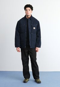 Marineblaues, knöpfbares Arbeitsjackett mit vier Fronttaschen und einem Logo-Aufnäher, kombiniert mit schwarzen Hosen und grünen Camouflage-Schuhen vor einem einfarbigen Hintergrund.