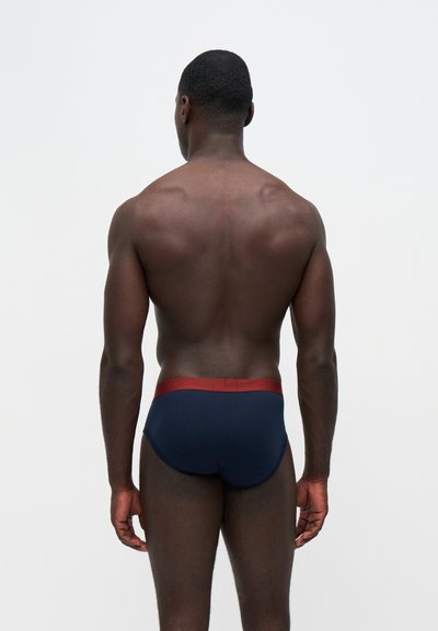 Homme vu de dos portant un slip bleu marine avec une ceinture rouge sur un fond clair uni.