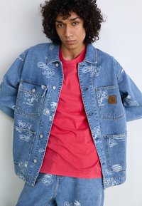 Denim jacket featuring various printed logos in white, a chest pocket, and a brown label patch, paired with a red crew-neck t-shirt.