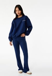 Sudadera azul marino con bolsillo y texto "marcador de tendencias". Pantalones a juego con franjas laterales y diseño acampanado. El material parece suave y liso.