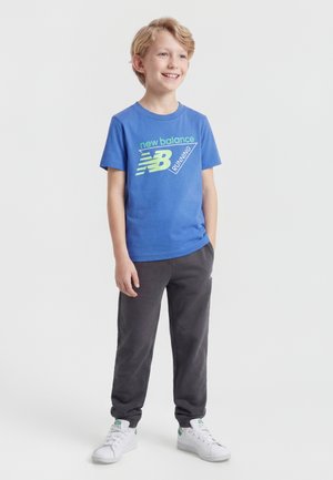Uśmiechnięty blond chłopiec stoi, ubrany w niebieską koszulkę do biegania New Balance, ciemne spodnie i białe trampki na jednolitym białym tle.