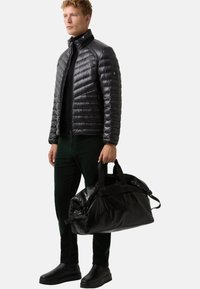 Schwarze Puffjacke mit horizontaler Steppung, Reißverschluss und hohem Kragen; kombiniert mit dunklen grünen Hosen und einer großen schwarzen Sporttasche.