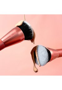 To vinklet makeup-børster med sorte og hvide børster holder flydende foundation, der drypper mod en blød lyserød baggrund.