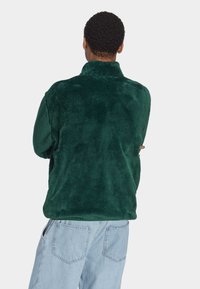 Sudadera de forro polar verde oscuro con cuello alto y superficie suave y texturizada, combinada con vaqueros azul claro anchos con bolsillos traseros.
