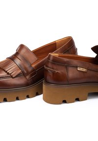 Mocasines de cuero marrón con acentos de flecos, punta redondeada y suela de goma gruesa con un patrón de relieve. Etiqueta de la marca visible en el lateral.