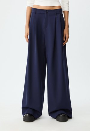 Vrouw draagt een high-waist, wijd uitlopende marineblauwe broek gecombineerd met zwarte schoenen en een cropped wit topje, staande tegen een effen achtergrond.