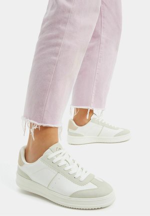 Pieds portant des baskets blanches et gris clair associées à un jean rose clair à ourlet effiloché sur un fond blanc uni.