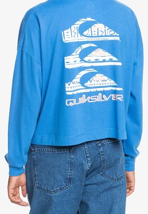 Osoba ubrana w niebieską koszulę z długim rękawem z białym wzorem logo Quiksilver na plecach oraz niebieskie dżinsy, widziana od tyłu.