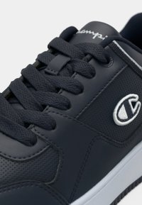 Granatowy sneaker z cholewką wykonaną ze skóry, perforowanymi elementami, płaskimi sznurowadłami oraz detalem z logo. Biała gumowa podeszwa i kontrastowe przeszycia jako akcenty.