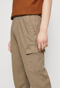 Khaki cargo kalhoty s texturovanou tkaninou, elastickým pasem, bočními kapsami a klopovou kapsou na stehně. Představují uvolněný střih.
