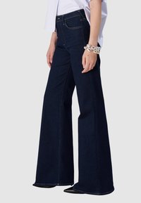 Femme portant un jean large bleu foncé, un haut blanc, des bracelets métalliques imposants et des chaussures noires à bouts pointus sur fond uni.