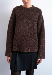 Femme portant un pull à manches longues marron texturé et une jupe plissée noire, debout devant un fond gris clair uni.