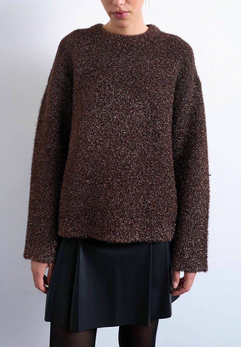 Femme portant un pull à manches longues marron texturé et une jupe plissée noire, debout devant un fond gris clair uni.