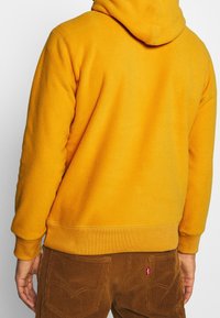 Sudadera con capucha de forro polar amarilla con bolsillo canguro y capucha ajustable. Puños y borde inferior acanalados, combinada con pantalones de pana marrones con bolsillos traseros.