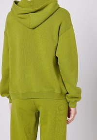 Felpa verde oliva con un tessuto a maglia testurizzato, taglio oversize, collo a cappuccio e polsini a costine; abbinata a pantaloni coordinati nello stesso materiale.
