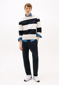Felpa a righe bianco e blu navy con scollatura a girocollo, indossata sopra una camicia di denim blu. Pantaloni neri e sneakers bianche completano l'outfit.