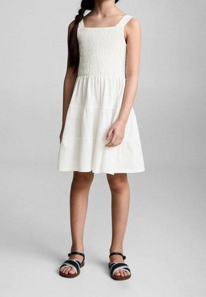 Dziewczyna w białej, bezrękawnikowej sukience z marszczonym gorsetem i falbanami na spódnicy, stojąca boso na neutralnym tle.