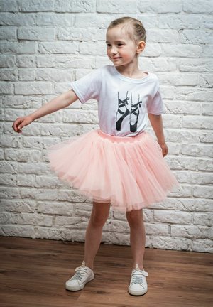 Weißes T-Shirt mit schwarzem Ballettschuh-Graphic, kombiniert mit einem flauschigen rosa Tüllrock und weißen Sneakern an einem Kind, das vor einer Mauer aus Ziegelsteinen steht.