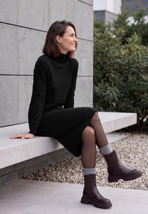 Une femme souriante est assise sur un banc en pierre à l’extérieur. Elle porte une robe noire, des chaussettes grises et des bottines marron foncé.
