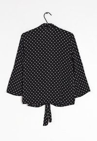 Svart topp med vita prickar, långa ärmar, knuten vid midjan, slät textur, avslappnad passform och krage.