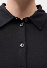 Gros plan sur une chemise noire à col avec quatre boutons gris fermés, portée sur le cou et le haut de la poitrine d'une personne.