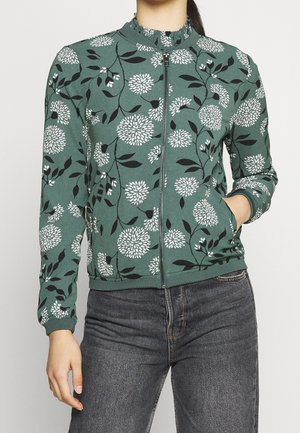 Grüne Jacke mit Reißverschluss und weißem Blumenmuster sowie schwarzen Blättern, getragen mit dunkelgrauen Jeans, eine Hand in der Tasche, Oberkörper sichtbar.