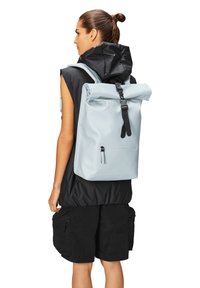 Mochila de rollo azul claro con herrajes negros, material texturizado, bolsillo frontal y correas ajustables, llevada por un modelo con un atuendo negro.