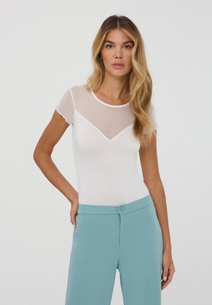 Top bianco aderente con maniche corte, caratterizzato da un pannello in rete trasparente sul petto, abbinato a pantaloni leggeri azzurri a vita alta.