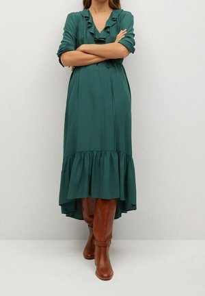 Femme portant une robe midi verte à manches longues avec des détails volantés et des bottes en cuir marron montant jusqu'aux genoux, debout, les bras croisés.