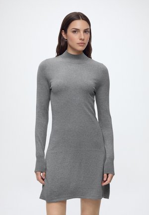 Femme aux longs cheveux bruns portant une robe en maille grise ajustée à manches longues et col montant, debout devant un fond clair uni.