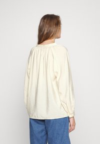 Blusa leve de tom creme, com decote franzido, mangas longas e um corte relaxado, combinada com calças de ganga azuis.