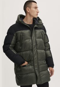 Polstret parkas i mørkegrønn og svart, med hette, sidelommer og quiltet tekstur. Behagelig lengde med glidelås foran.