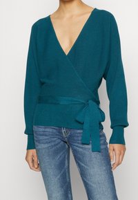 Teal wikkeltrui in ribbelstructuur met een diepe V-hals en lange mouwen, voorzien van een gestrikt taille-detail. Gecombineerd met blauwe denim jeans.