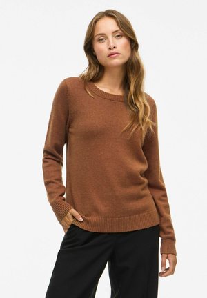 Femme aux longs cheveux châtain clair portant un pull marron à manches longues et un pantalon noir, debout avec une main dans la poche sur un fond uni.