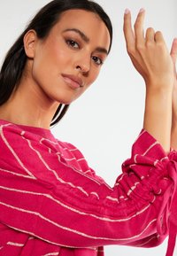 Yksi fuksia raidoitettu yläosa, jossa on metalliset kultaraitat, varustettuna kerrotuilla hihoilla ja nauhoilla. Kankaalla on pehmeä tekstuuri, joka on suunniteltu rentoa ilmettä varten.