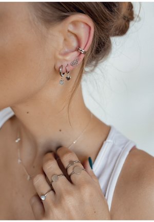 Silberohrringe umfassen Ohrringe mit Charms: ein Herz und eine Blume; ein Ohrcuff mit Blattdesign. Ringtragende Finger zeigen schlichte Silberbänder.