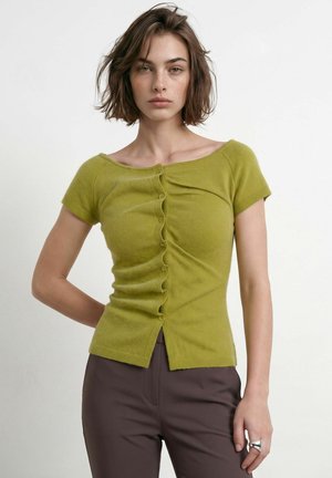 Femme aux cheveux courts et bruns portant un haut boutonné vert olive ajusté et un pantalon brun foncé, debout avec une main derrière le dos.