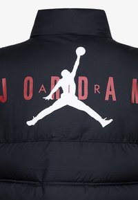 Chaqueta de plumas negra con un gran logo blanco de Jordan y texto rojo que dice "J O R D A N" en la parte de atrás, con un cuello alto.