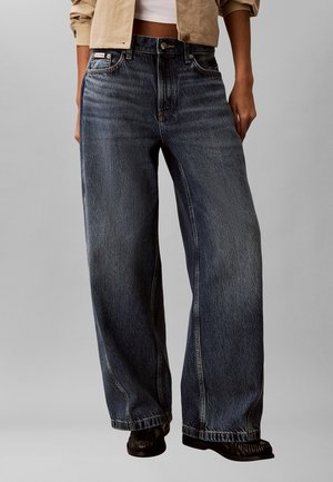 Person trägt dunkelblaue Jeans mit hoher Taille und weitem Bein sowie schwarze Schuhe, kombiniert mit einer beigen kurzgeschnittenen Jacke und weißem Oberteil.
