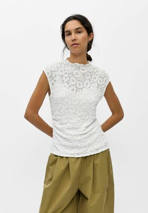 Femme aux cheveux foncés attachés porte un haut en dentelle blanc sans manches et un pantalon ample vert olive, debout les mains dans le dos.