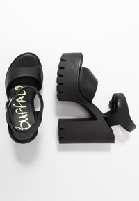 Sandalias negras de tacón alto con plataforma gruesa, hechas de material sintético texturizado. Cuentan con una correa ajustable en el tobillo y un logo en la plantilla.