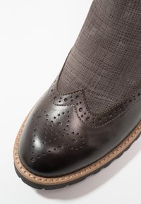 Gros plan sur l’avant d’une chaussure en cuir brun foncé avec perforations brogue, surpiqûres couleur tan contrastantes et dessus en tissu texturé.