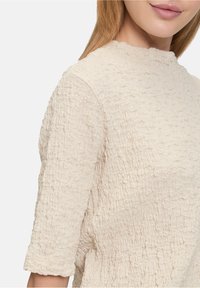 Blouse crème texturée avec un col haut et des manches trois-quarts. Le tissu présente un design froissé, ajoutant de la profondeur et de la dimension.