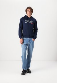 Marineblauer Hoodie mit weißem und rotem Logo, Kängurutasche, kombiniert mit hellblauen Jeans und schwarzen Schuhen. Einfaches Design, lässige Passform.