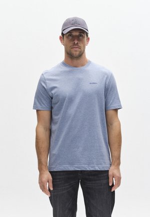 Bărbat în picioare, purtând un tricou Gabba bleu deschis, blugi închiși la culoare și o șapcă gri, pe un fundal alb simplu.